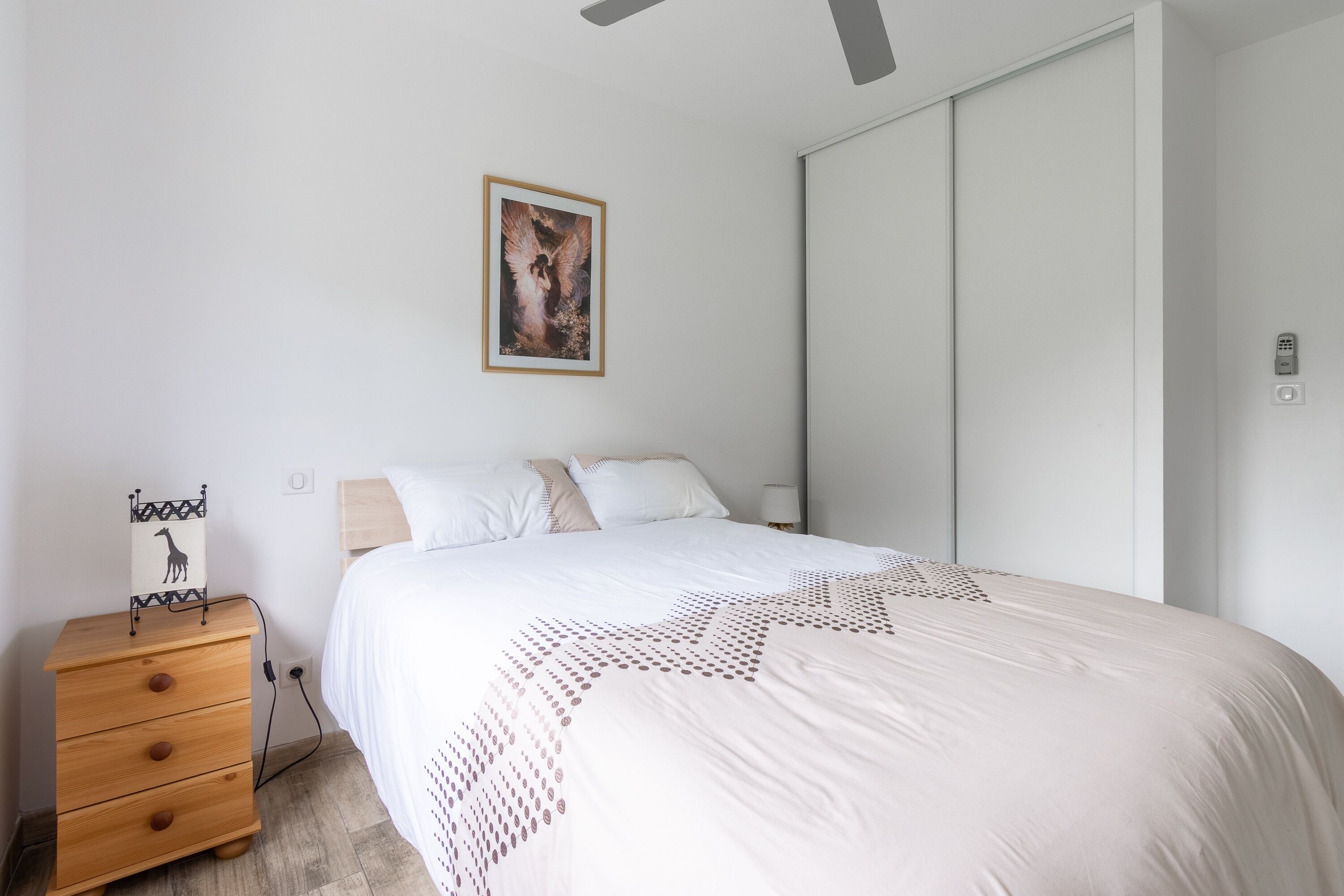 A bedroom with a bed, a wooden dresser, a nightstand, a wall-mounted light, and a framed picture.
