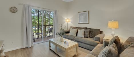 A living room with a sofa, a coffee table, a lamp, a ceiling fan, a clock, and a view through a sliding glass door.