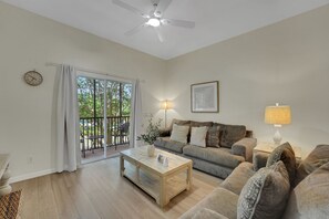 Ein Wohnzimmer mit einer Couch, einem Couchtisch, einer Lampe, einem Deckenventilator, einer Uhr und Blick durch eine Glastür.