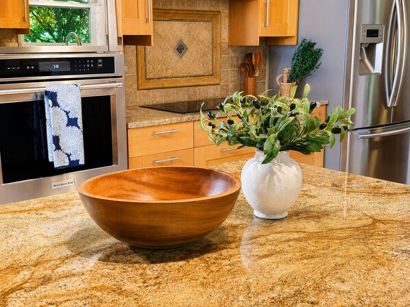 A kitchen with a granite countertop, a wooden bowl, a white vase with green plants, a stainless steel refrigerator, and a built-in oven.