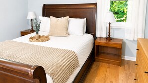 Un lit bien fait, avec une tête de lit en bois, deux tables de chevet avec des lampes, une fenêtre avec des rideaux et une petite plante posée sur la table de chevet.