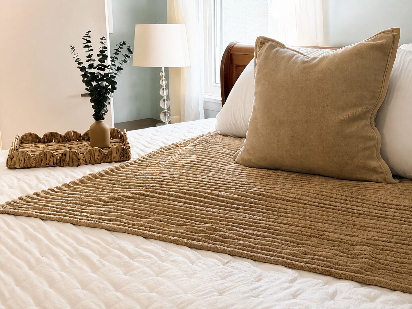 A neatly made bed with a beige blanket, a white pillow, and a decorative woven tray.