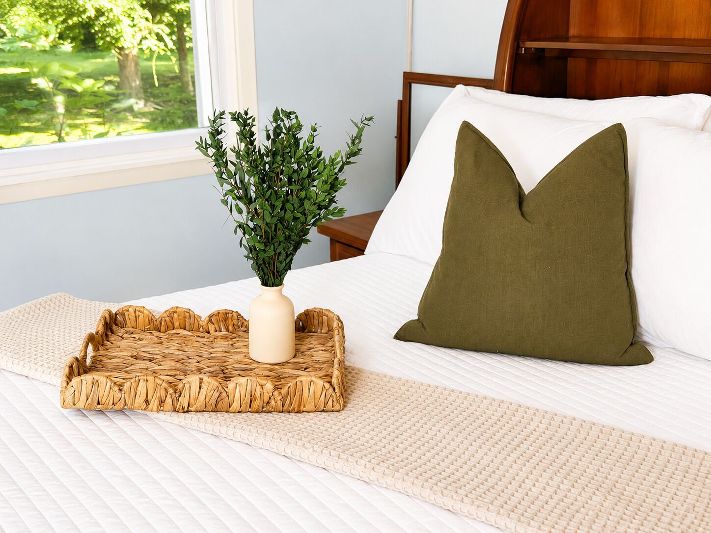 A neatly made bed with a white comforter, a green pillow, a woven tray, and a vase with green foliage.