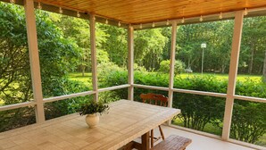 Une table en bois, un banc, une plante en pot et une vue sur un jardin luxuriant, visible à travers de grandes fenêtres.