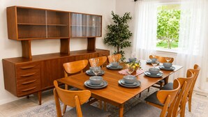 Une salle à manger avec une table en bois dressée pour six personnes, entourée de six chaises, un buffet en bois avec des portes en verre et une plante en pot.