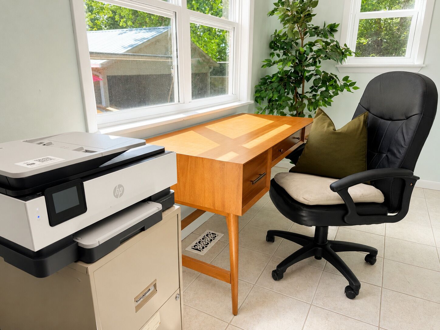 A home office with a printer, wooden desk, black office chair, and a potted plant.