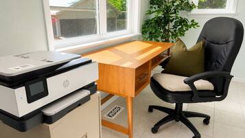 A home office with a printer, wooden desk, black office chair, and a potted plant.
