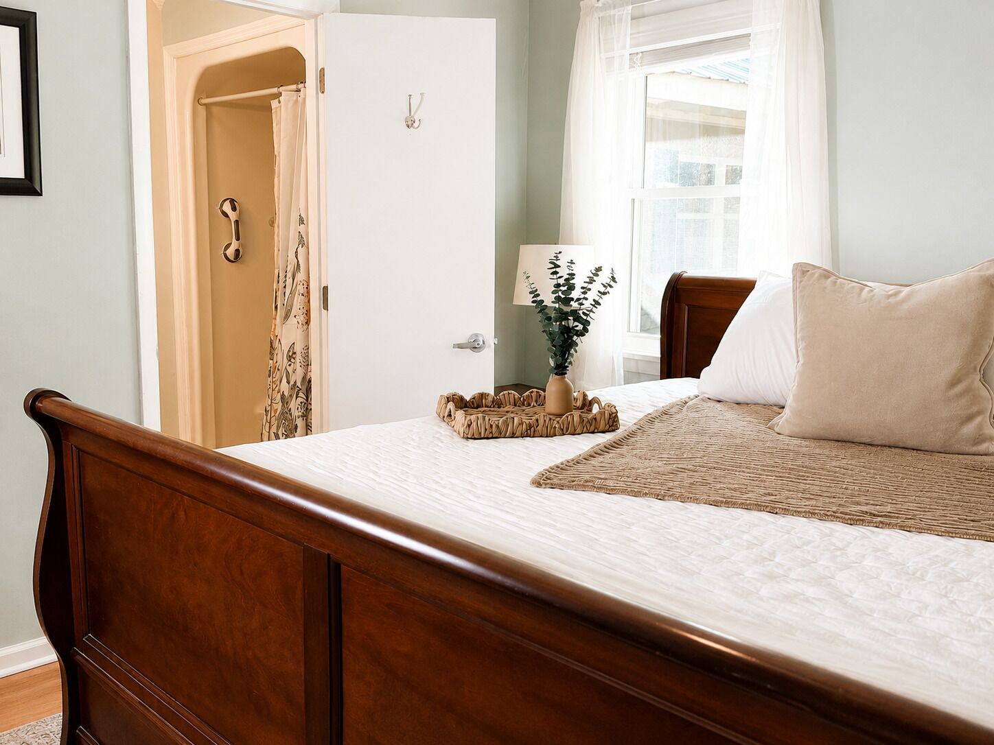 Un dormitorio con una cama de madera, una mesita de noche con una lámpara y una planta, una puerta con cortina y una ventana con cortinas de encaje.