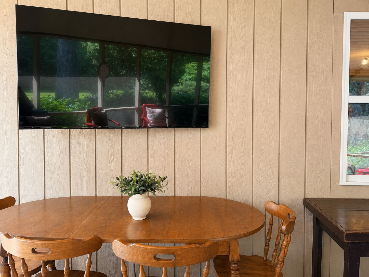 Un comedor con una mesa redonda de madera, sillas, un florero con planta y un televisor montado.