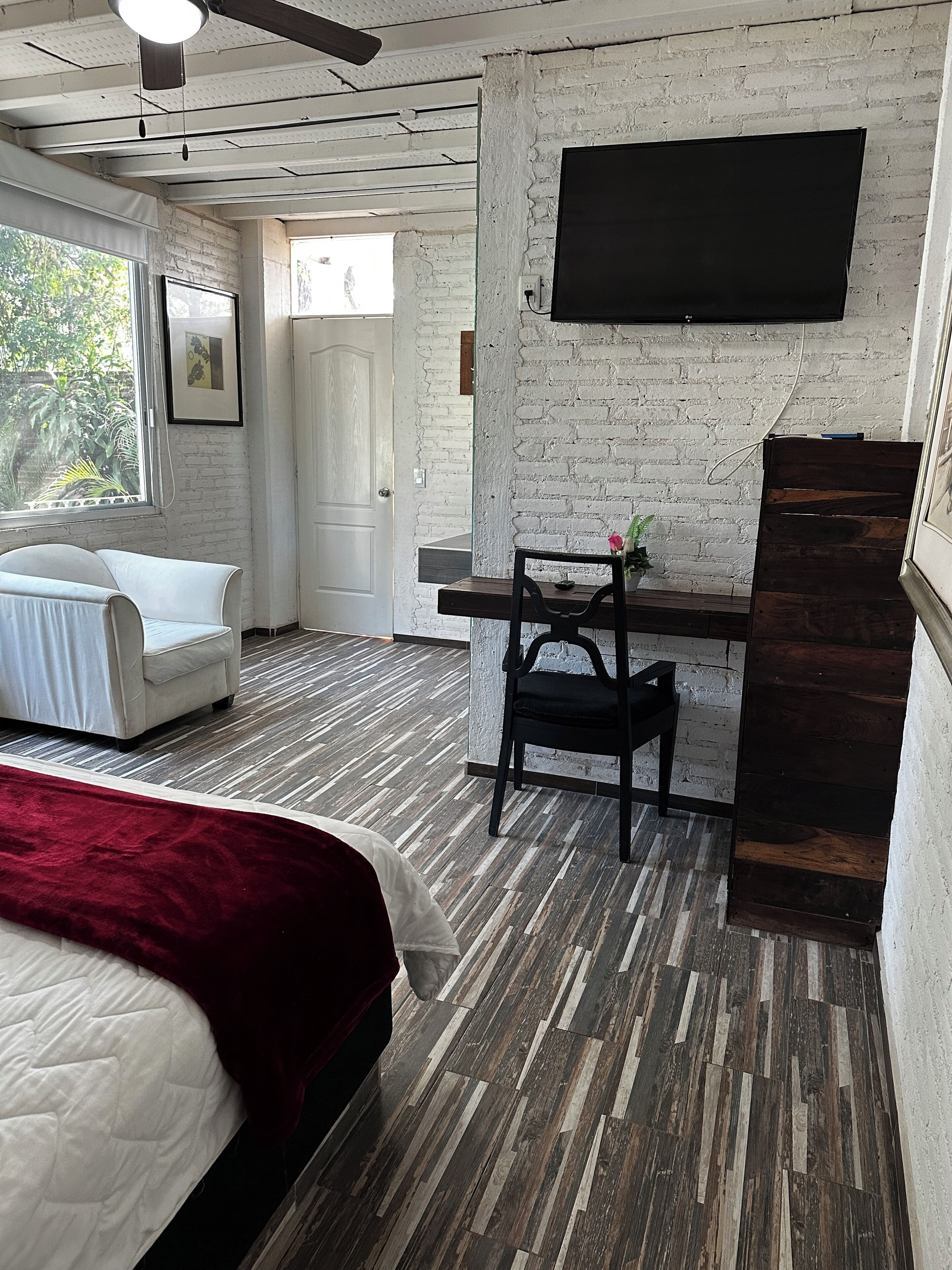 Een kamer met een bed, een bureau, een stoel, een aan een bakstenen muur bevestigde televisie en een raam met uitzicht op groen.