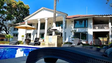 Un área de piscina con sillones reclinables, una estatua y un edificio con columnas y tejado de tejas rojas.