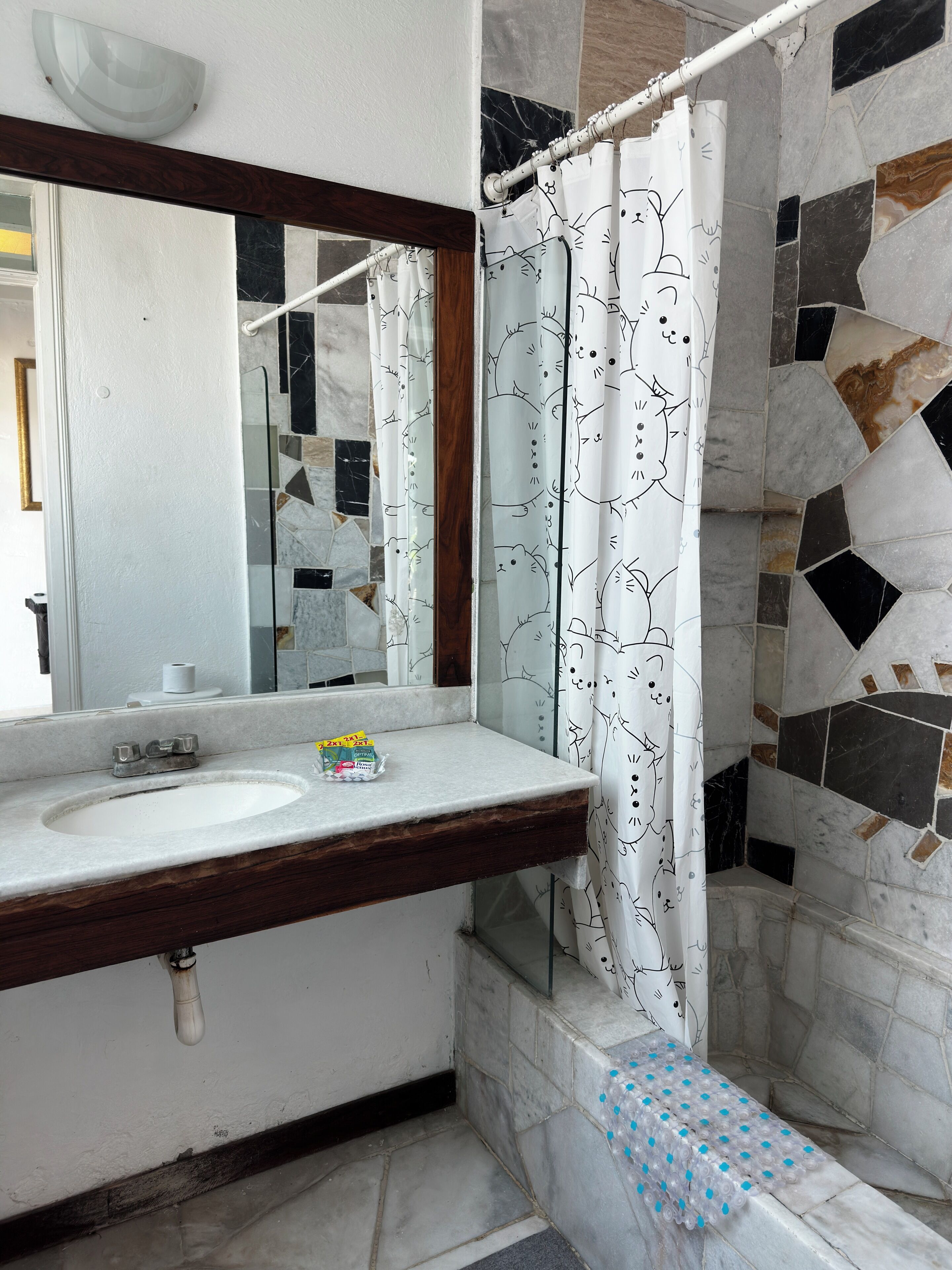 Un baño con un lavamanos blanco, un espejo, una cortina de ducha con estampado de gato y una pared de mosaicos.