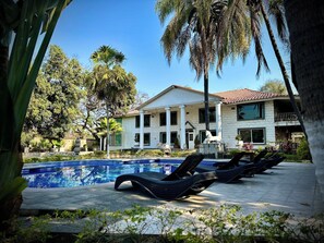 A pool area with lounge chairs, a white building with columns, and palm trees. - Casa vintage & eventos  (Yautepec)