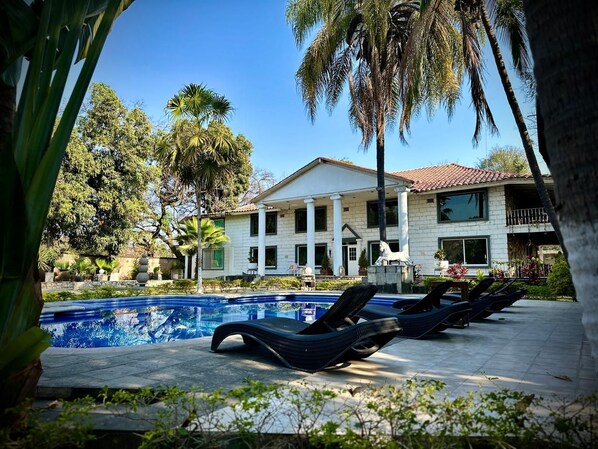 Un área de piscina con sillones reclinables, un edificio con columnas y palmeras.