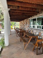 An outdoor patio with wooden tables and chairs, stone walls, and a view of the surrounding garden.
