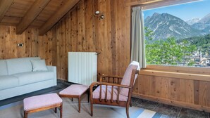 Interior de una cabaña de madera con sofá, dos sillones, un radiador y una ventana con vistas a la montaña.