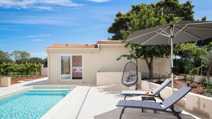 Una piscina con sillones de descanso y una hamaca colgante, un edificio pequeño con puerta corrediza y un árbol al fondo.
