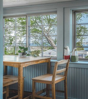 A dining area with a wooden table and chairs, large windows with a view of trees and a house, and a potted plant on the windowsill. - Stay at Back Beach Rockport (Rockport)
