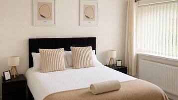 A neatly made bed with a black headboard, two framed artworks on the wall, and a window with blinds.
