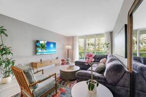 A modern living room with a sofa, armchair, coffee table, and a TV on the wall. - Walk to Ocean | Luxury 2BR w/ King Master & Pool (Sunny Isles Beach)