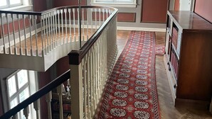 A spacious room with a wooden floor, a red patterned carpet, a white railing, and a large window. - Historic Copenhagen Villa with Garden (København)