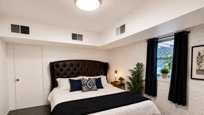 Ein Schlafzimmer mit einem großen Bett, einer weißen Tür, einem Fenster mit Vorhängen und einer Pflanze auf der Fensterbank.