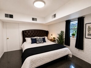 Ein Schlafzimmer mit einem großen Bett, einer weißen Tür, einem Fenster mit Vorhängen und einer Pflanze auf der Fensterbank.
