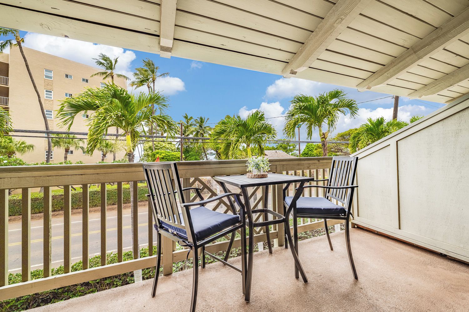 A covered outdoor seating area with a table and chairs, overlooking a street with palm trees and buildings.