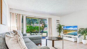 Un soggiorno con divano, tavolino, televisore e vista su un giardino attraverso ampie finestre.
