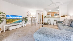 Un soggiorno moderno con una TV a schermo piatto, una zona pranzo con sgabelli, una cucina con mobili in legno e una ventola a soffitto.