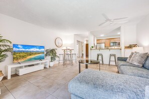 A modern living room with a flat-screen TV, a dining area with stools, a kitchen with wooden cabinets, and a ceiling fan. - Kihei Garden Estates | King Bed, Pool, Hot Tub, Private Lanai & Steps to Beach (Kihei)