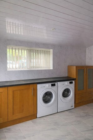 Ein Waschraum mit zwei Waschmaschinen, die von vorne geladen werden, Holzausbau, einem Fenster mit Jalousien und einem gefliesten Boden.