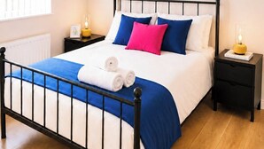 Una cama bien hecha con colcha azul y blanca, almohadas en azul y rosa, y toallas enrolladas al lado. Hay obras de arte enmarcadas en la pared sobre la cama.