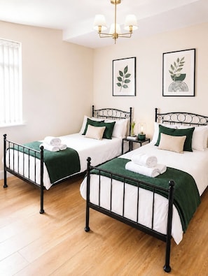 Ein Hotelzimmer mit zwei Betten, jeweils mit grüner Bettdecke und weißen Laken, einem schwarzen Metall-Kopfschild und einer wandmontierten Lampe.