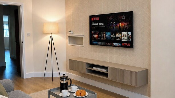 Una sala de estar moderna con un televisor de pantalla plana montado en la pared, una mesa de centro con una cafetera y pasteles, un sofá gris y una lámpara.