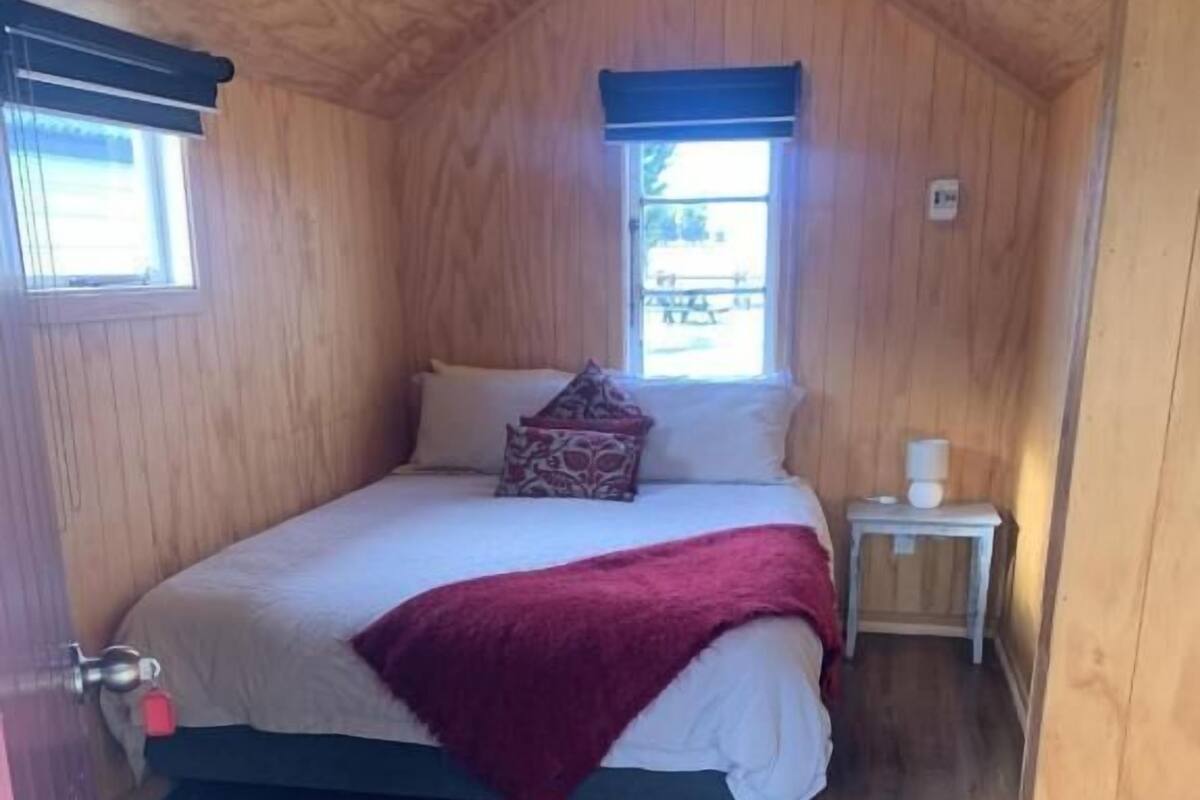 A bedroom with a wooden ceiling, a bed with a red blanket, a nightstand, and a window.