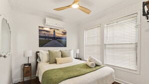 Una camera da letto con un letto, un comodino, una lampada, un quadro raffigurante un faro, una ventola a soffitto e persiane bianche.