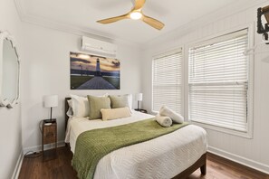 Quarto com cama, mesa de cabeceira, luminária, pintura de um farol, ventilador de teto e persianas brancas.