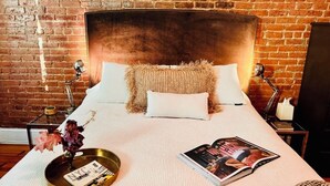 A bed with a brick wall backdrop, a bedside lamp, a magazine, and a tray with a book and flowers. - Stay with a local in Brownstone Brooklyn's Carroll Gardens (Brooklyn)