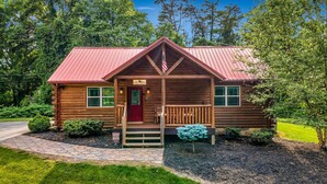A log cabin with a red roof, a porch with a bench, and a small American flag. - Dollywood Escape | Hot Tub • Fire Pit • Arcade (Pigeon Forge)