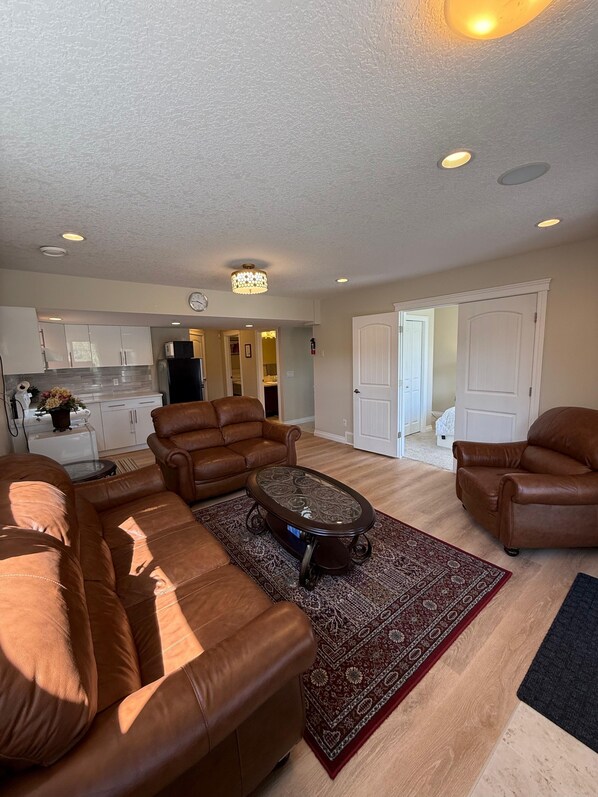 A living room with a sectional sofa, a coffee table, and a patterned rug. There is a kitchen in the background with white cabinets and a refrigerator.