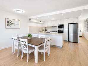 A modern kitchen with white cabinetry, a central island, and a dining area with white chairs. - Relax & Unwind 2BR w/ Parking Maroochydore Beach (Sunshine Coast)