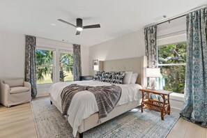 Ein Schlafzimmer mit einem Bett, einem Deckenventilator, einem Fenster mit Vorhängen, einem Sessel und Blick auf Grün.