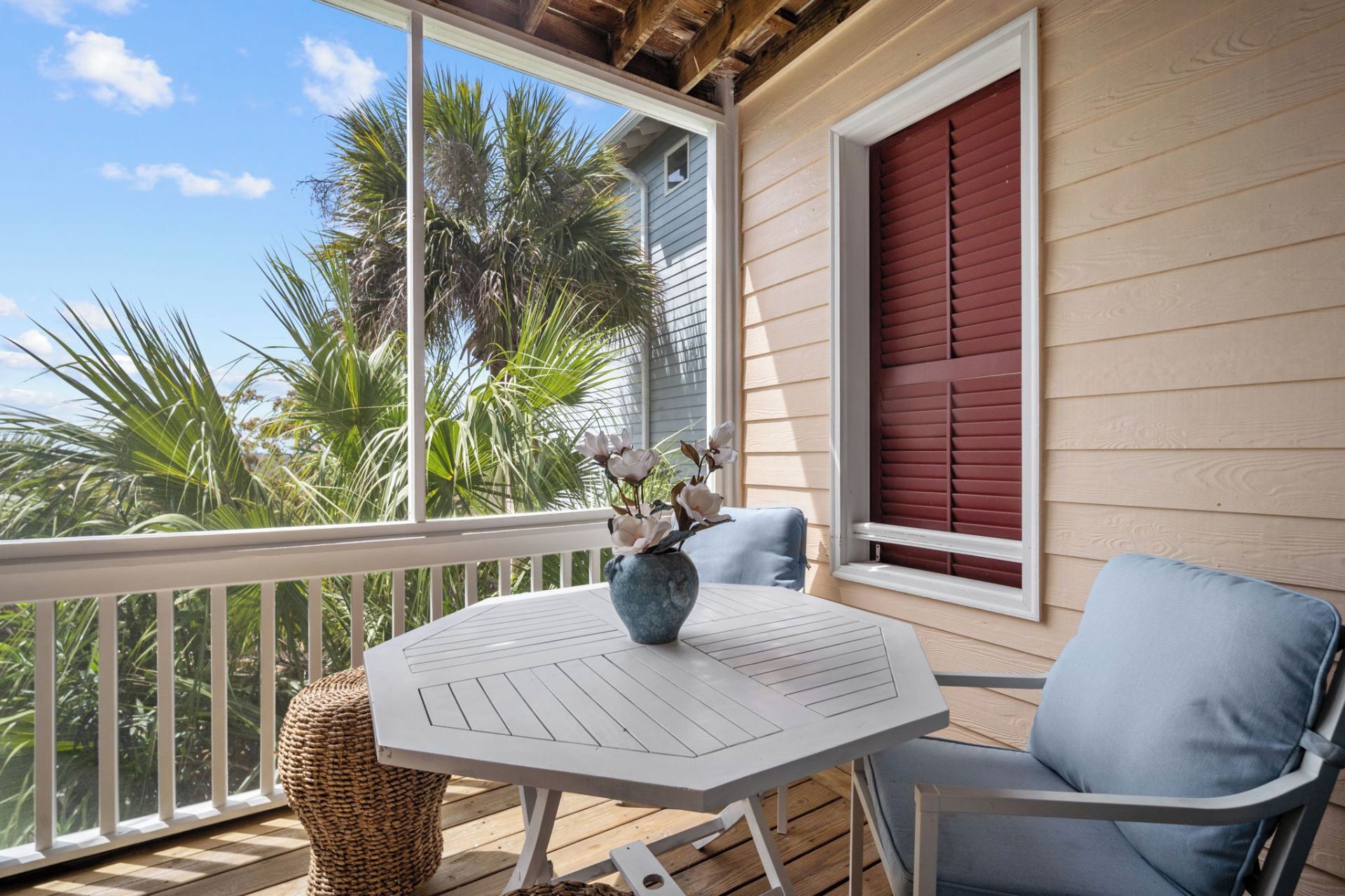 A balcony with a white table and blue chair, a vase with flowers, and a red window shutter.