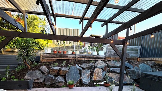 An outdoor patio with metal chairs, potted plants, a stone wall, and a pergola structure. - Near Movie World & Wet'n'Wild | 2BR Home | Parking (Oxenford)