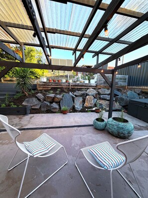En udendørs terrasse med metalstole, krukker med planter, en stenvæg og en pergola.