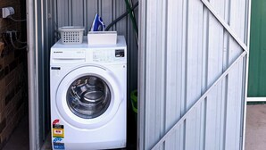 A compact laundry room with a front-loading washing machine, a drying rack, and a folding table. - Near Movie World & Wet'n'Wild | 2BR Home | Parking (Oxenford)