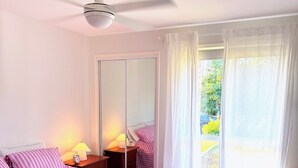 A bedroom with a bed, a ceiling fan, a nightstand, a lamp, a mirror, and a view of the outdoors. - Near Movie World & Wet'n'Wild | 2BR Home | Parking (Oxenford)