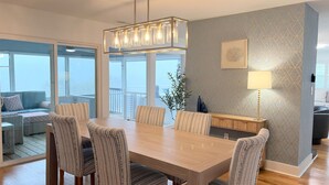 Een eetgedeelte met een houten tafel, gestreept beklede stoelen, een hanglamp, een bijzettafeltje met een lamp, en uitzicht door grote glazen deuren op een buitenbalkon.