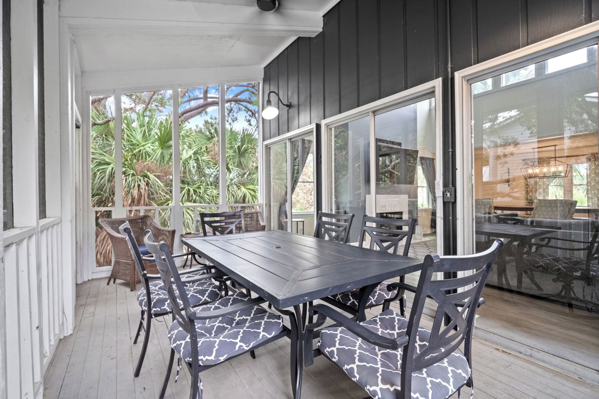 An outdoor patio with a wooden table and chairs, large windows, and a view of greenery.